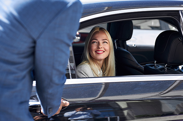 Eine hübsche blonde Frau lächelt einen Chauffeur an, der ihr die Wagentür öffnet.