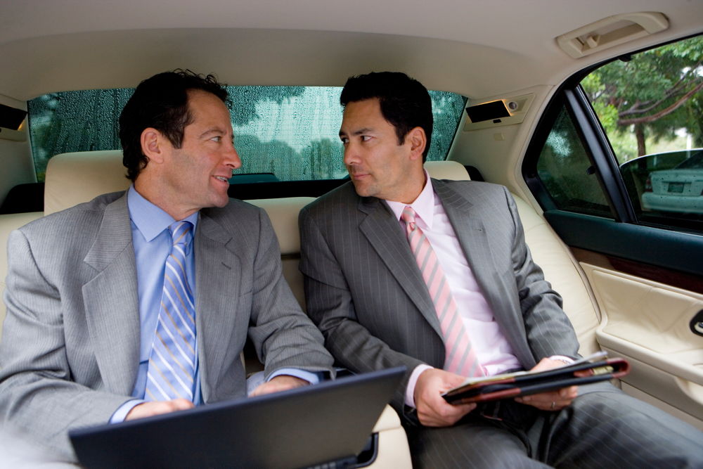 Zwei Gechäftsmänner in schicken Anzügen sitzen im Fond einer Chauffeur Limousine und lassen sich zu einem Geschäftstermin fahren.