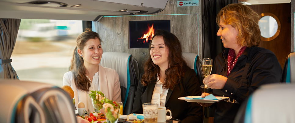 lounge-liner-bus-on-board-kitchen Two young women are being served food and drink in the Lounge Liner operated by German Limousines Network GmbH & Co. KG. This turns a trade fair visit with the whole company into an incentive trip.